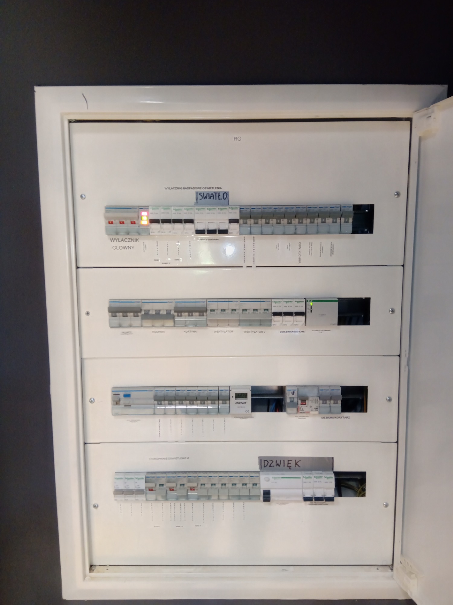 Otwarta, biała skrzynka rozdzielcza z wyłącznikami nadprądowymi i opisami obwodów, w tym 'Światło', 'Kuchnia', 'Wentylator', 'Dźwięk', z jednym wyłącznikiem w pozycji włączonej, wskazującym...