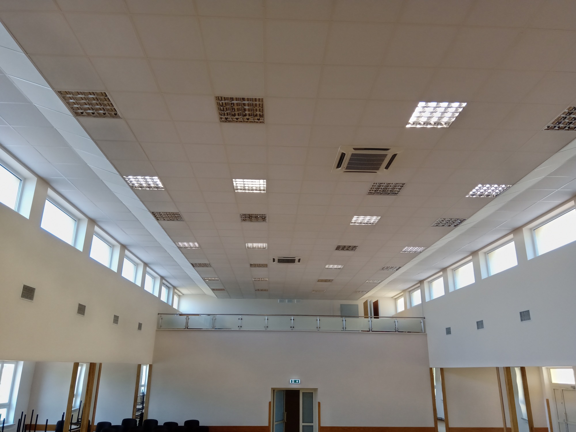 Wnętrze dużej sali z białym sufitem podwieszanym, oświetleniem rastrowym i klimatyzacją centralną, widok na antresolę z przeszkloną balustradą i rząd okien w górnej części ścian.