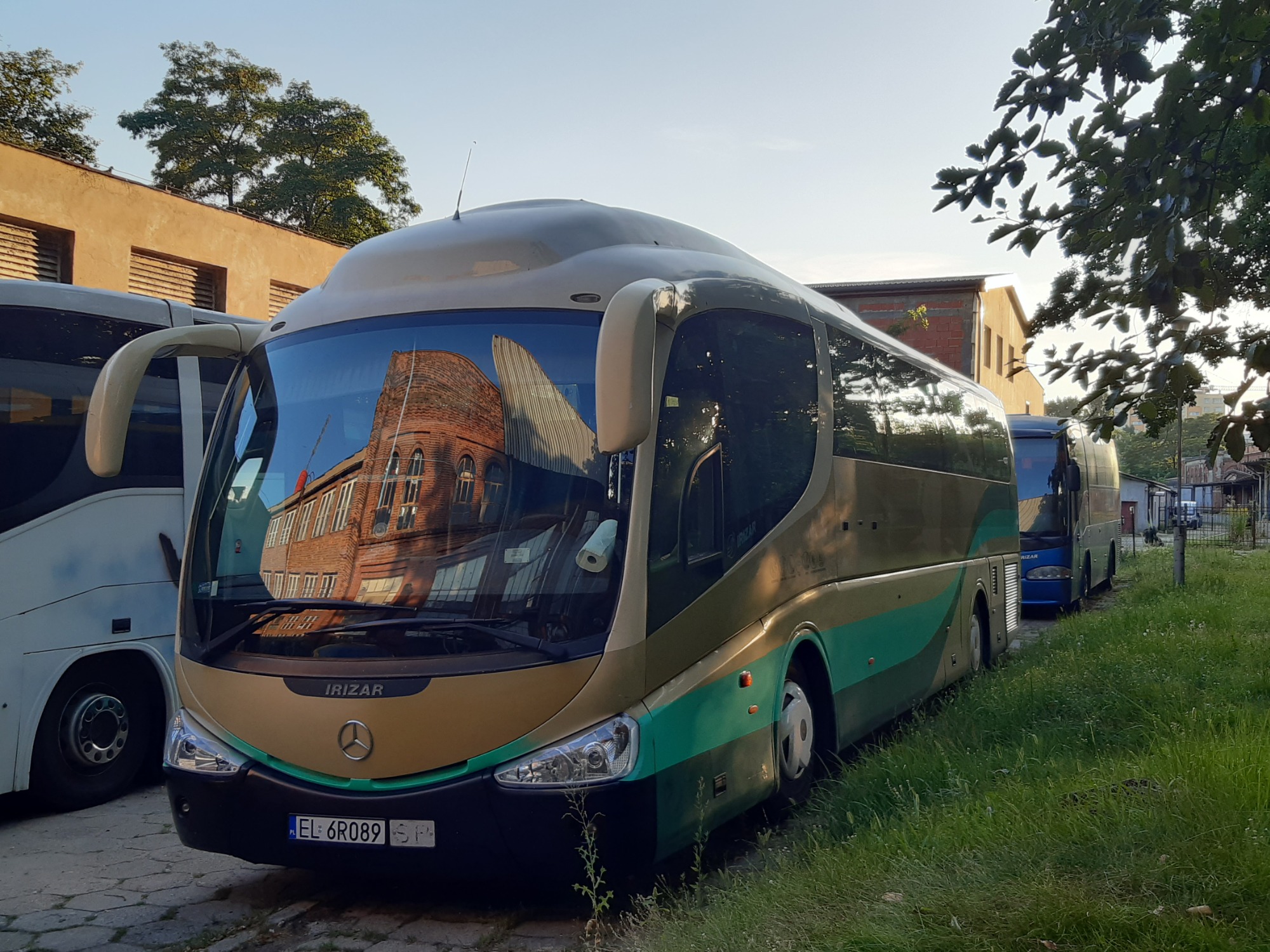 Autobus turystyczny Irizar w kolorach beżowo-zielonych, zaparkowany na placu z widoczną rejestracją EL 6R089, w szybie odbija się budynek z czerwonej cegły.