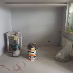 Pokój w trakcie remontu: świeżo pomalowane ściany w odcieniach szarości, zakryte folią okno, grzejnik, wiadro z farbą Dekoral, odkurzacz przemysłowy i zabezpieczone meble.