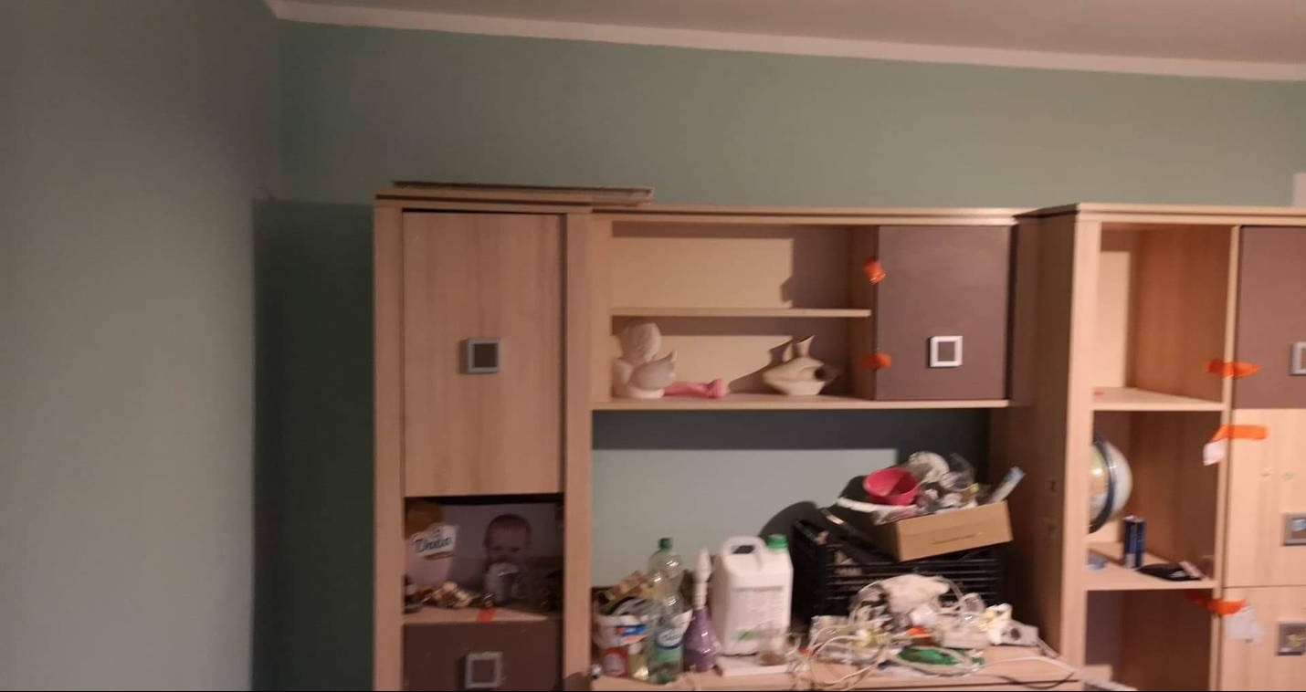 Pokój z zielonymi ścianami, umeblowany meblościanką z bałaganem na półkach i blacie, widoczne butelki, kartony, bibeloty i przewody.