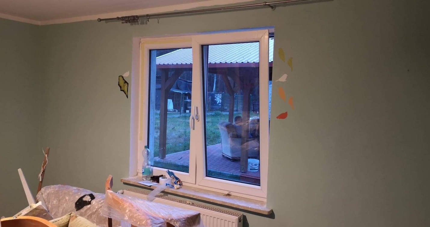 Pokój z pomalowanymi na zielono ścianami, z widocznymi śladami po taśmie malarskiej i naklejkami na ścianie, okno przesuwne z widokiem na altanę i meble ogrodowe, meble przykryte folią ochronną...