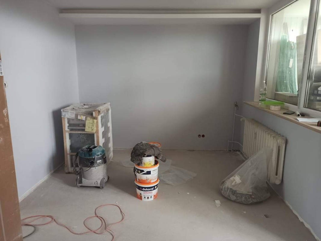 Pokój w trakcie remontu: świeżo pomalowane ściany w odcieniach szarości, zakryte folią okno, grzejnik, wiadro z farbą Dekoral, odkurzacz przemysłowy i zabezpieczone meble.