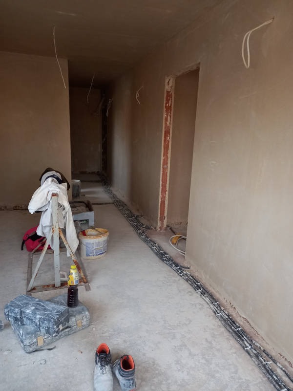 Wnętrze w trakcie prac wykończeniowych: ściany pokryte świeżo nałożoną gładzią, widoczne narzędzia, wiadro z farbą, kable elektryczne ułożone wzdłuż ściany, pomieszczenie w stanie surowym.