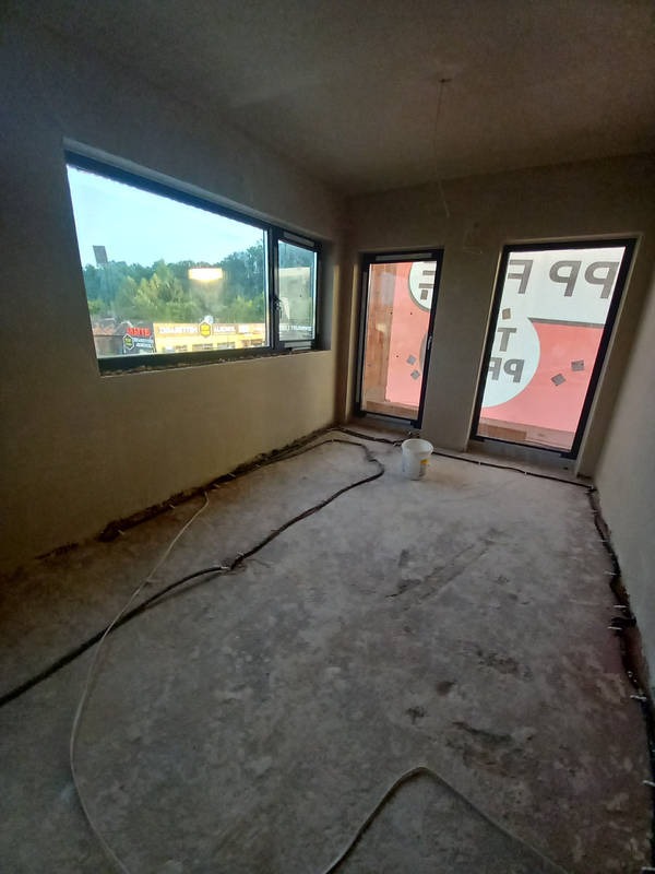 Surowe wnętrze pokoju z oknami i drzwiami balkonowymi, ściany przygotowane pod malowanie, widoczne przewody elektryczne na podłodze i wiadro po farbie, stan przed wykończeniem.