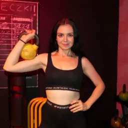 Uśmiechnięta kobieta w sportowym stroju pozuje z żółtym kettlebellem w siłowni, obok tablicy z rozpisanymi ćwiczeniami i stojaka z obciążeniami.