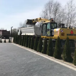 Żółta koparka Komatsu ładuje ziemię do wywrotki na tle rzędu tuj i kostki brukowej.