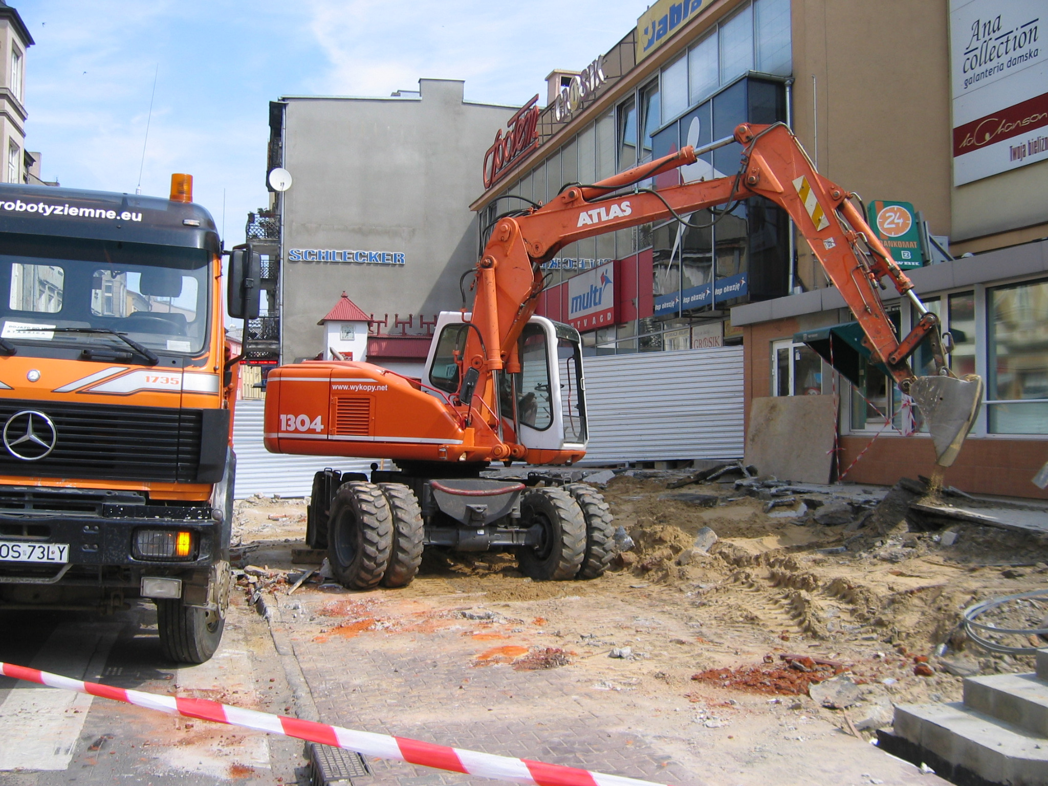 Pomarańczowa koparka Atlas na placu budowy w centrum miasta, obok stoi ciężarówka z napisem 'robotyziemne.eu', w tle budynki z reklamami.
