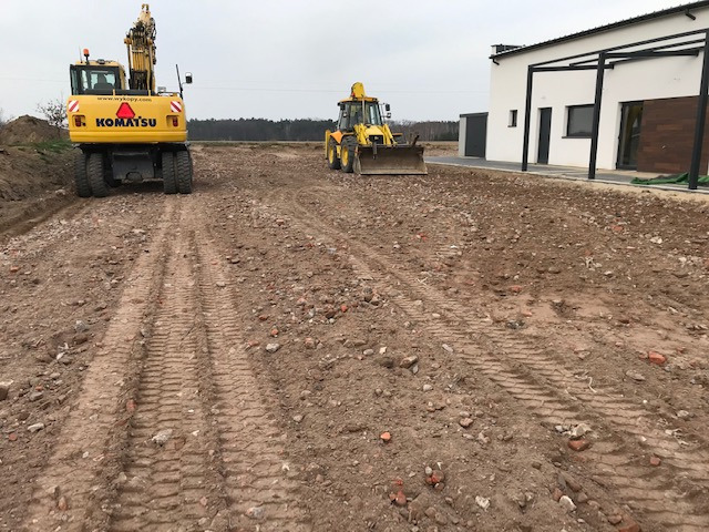 Żółta koparka Komatsu tyłem i traktor z przodu wyrównują teren pod budowę domu z pergolą, widoczne ślady opon na ziemi.