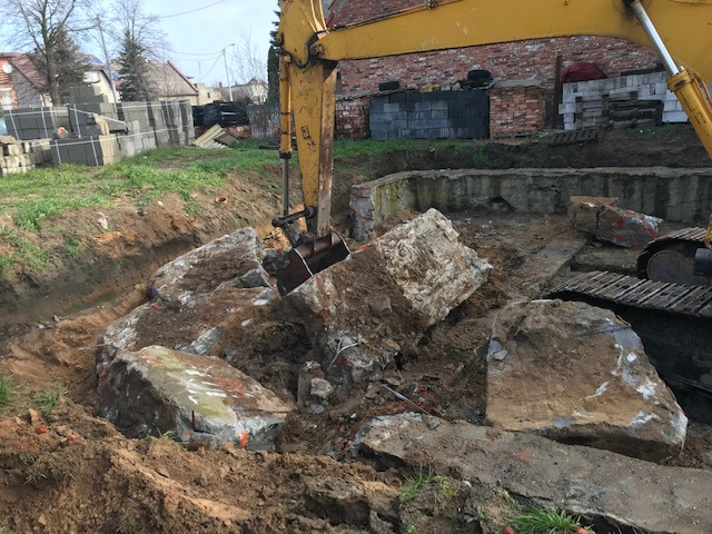 Żółta koparka z łyżką podnosi duże, szare bloki betonowe z wykopu, widoczne ślady po rozbiórce fundamentów i ceglany budynek w tle.