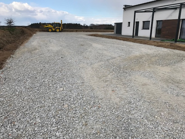 Teren po niwelacji z rozsypanym kruszywem, widoczny żółty traktor oraz budynek mieszkalny w tle.