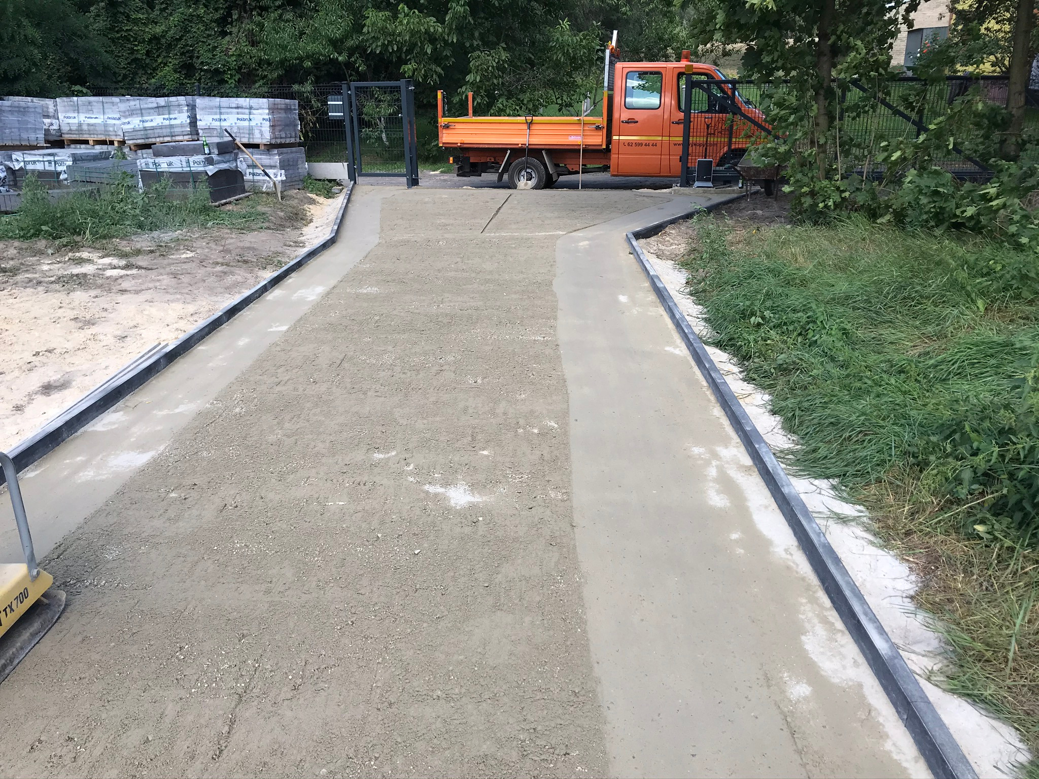 Świeżo wylana betonowa ścieżka z obrzeżami, częściowo jeszcze nieutwardzona, pomarańczowa wywrotka w tle, palety z kostką brukową po lewej stronie, zagęszczarka w lewym dolnym rogu.