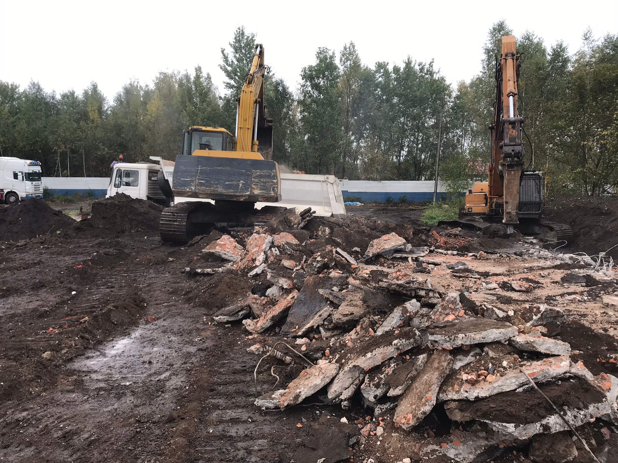 Dwa żółte koparki na placu budowy z gruzem i błotem, jedna z łyżką ładuje gruz do białej ciężarówki, w tle drzewa.