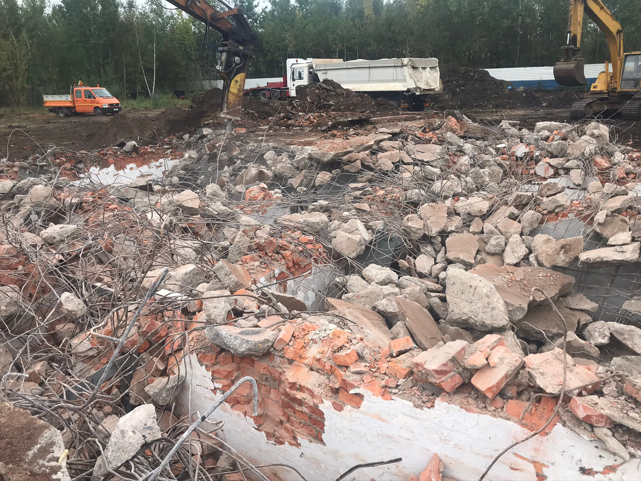 Gruzowisko z fragmentami cegieł i betonu po rozbiórce, z widoczną armaturą, na tle pracujących maszyn budowlanych i ciężarówki wywożącej urobek.