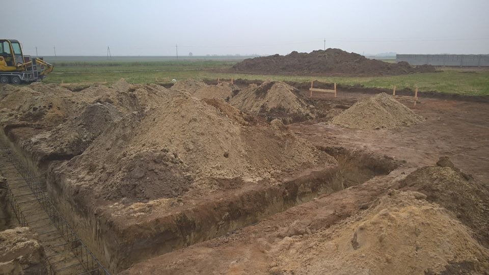 Przygotowany wykop pod fundamenty z widocznym zbrojeniem, usypane pryzmy ziemi i koparka w tle na rozległym terenie budowy.