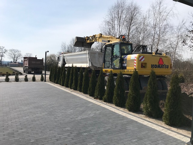 Żółta koparka Komatsu ładuje ziemię do wywrotki na tle rzędu tuj i kostki brukowej.