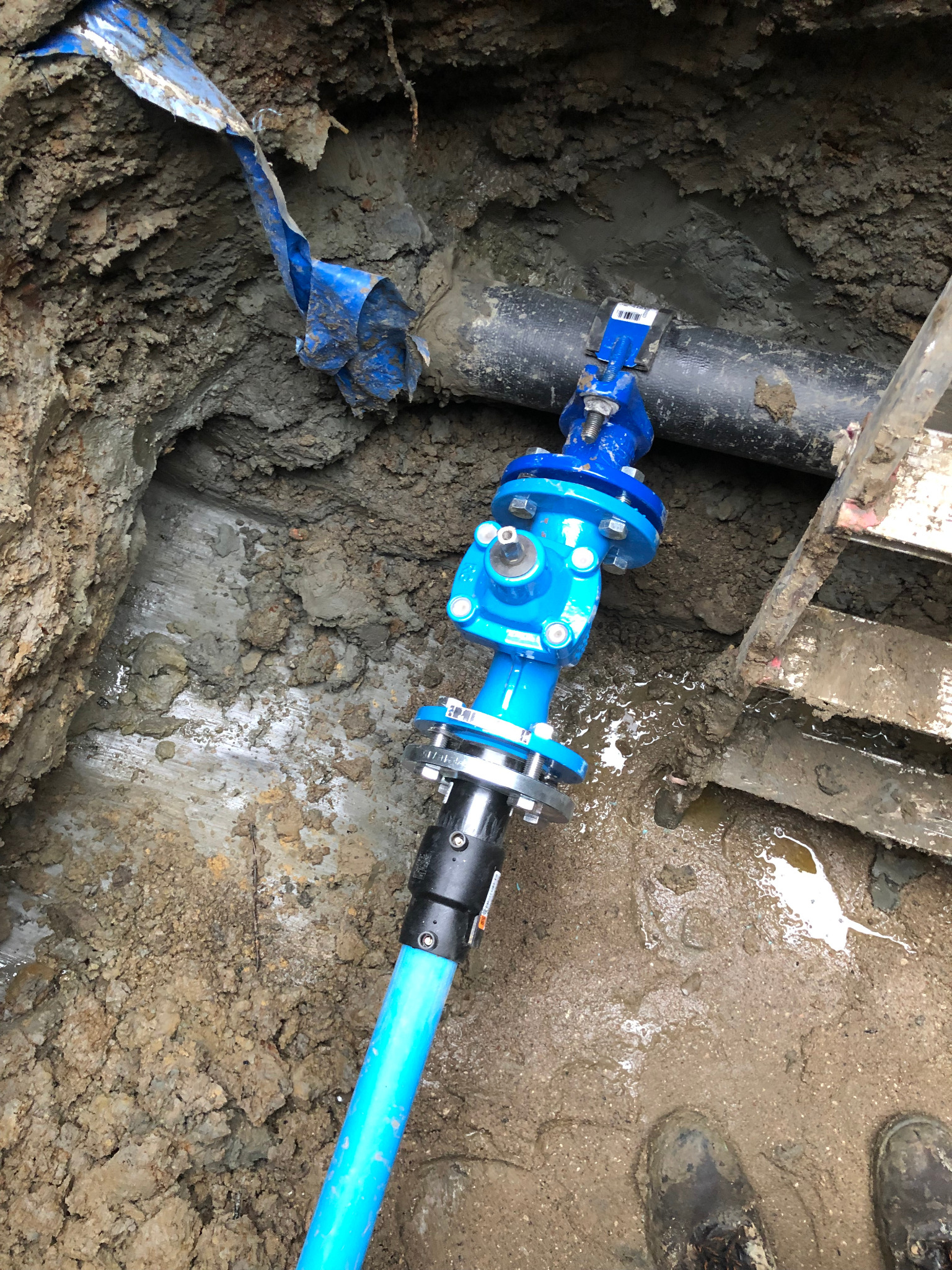 Niebieski zawór z rurą podłączony do czarnej rury w wykopie ziemnym, widoczny fragment drabiny i buty pracownika.