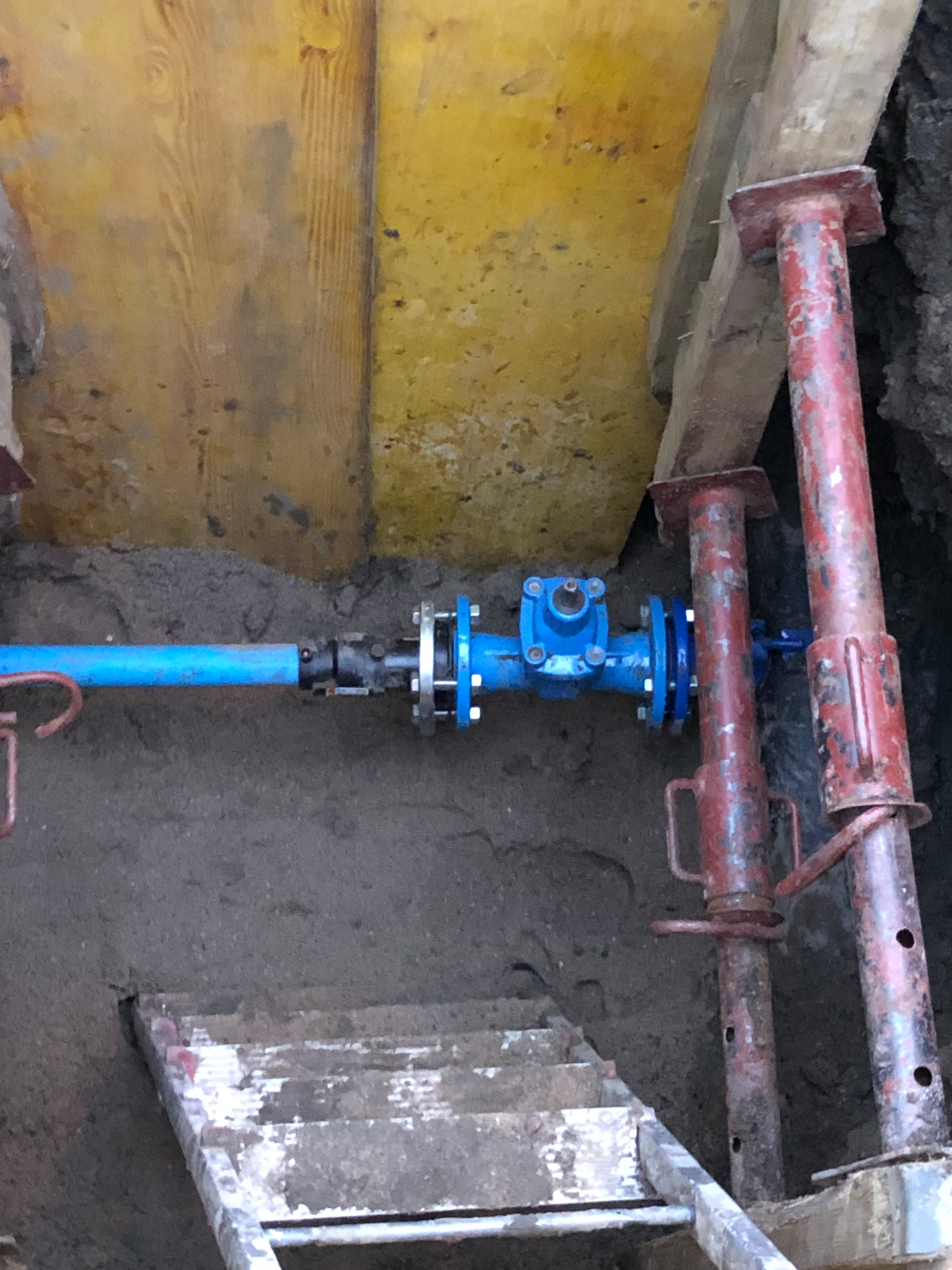 Niebieska rura wodociągowa z zaworem i kołnierzem, łączona w wykopie, widoczne stemple stalowe i drabina ze śladami zaprawy.