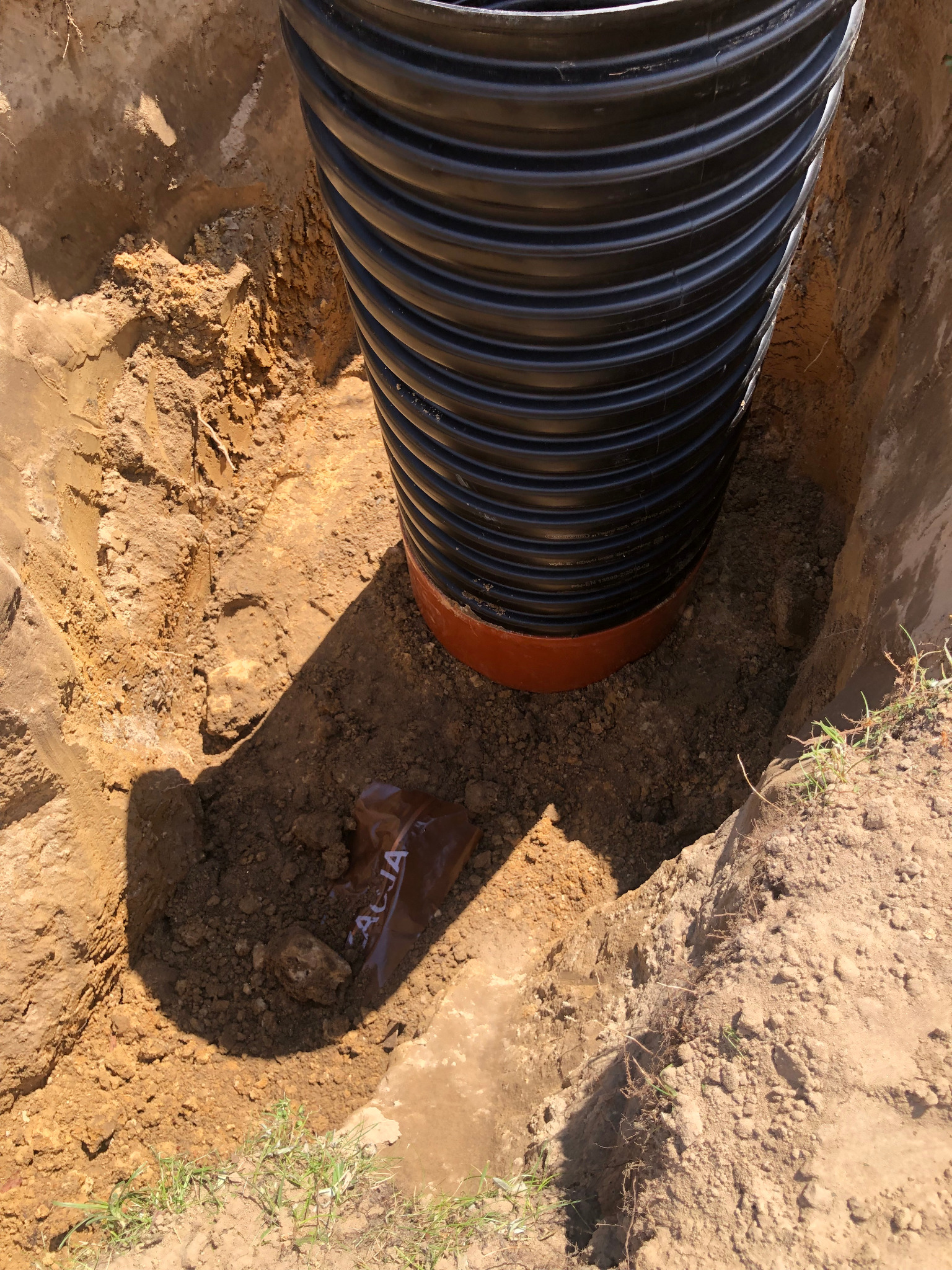 Czarna, karbowana rura w wykopie z ziemi, widoczna brązowa uszczelka i fragment foliowego opakowania leżącego na dnie.