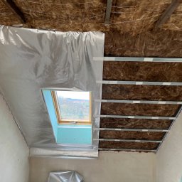 MBB Mateusz Bochorodycz Budownictwo - Skośny sufit poddasza w trakcie izolacji: wełna mineralna, folia aluminiowa i metalowy stelaż. Widoczne okno dachowe i fragment ściany. Prace wykończeniowe.