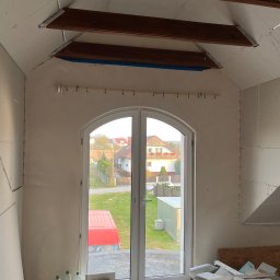 MBB Mateusz Bochorodycz Budownictwo - Poddasze w trakcie adaptacji: widoczne płyty gipsowo-kartonowe na ścianach i suficie, drewniane belki konstrukcyjne, okno balkonowe z widokiem na zewnątrz. Prace wykończeniowe w toku.