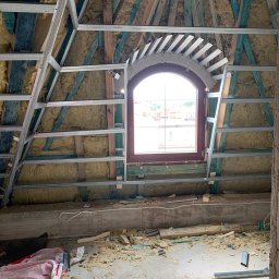 MBB Mateusz Bochorodycz Budownictwo - Poddasze w trakcie izolacji: wełna mineralna między krokwiami, metalowy stelaż pod płytę G-K, okno łukowe. Widoczne resztki materiałów na podłodze.