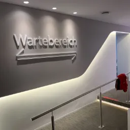 Korytarz biurowy z napisem 'Wartebereich' i strzałką wskazującą kierunek, oświetlony punktowo i listwami LED, z metalową barierką i czerwonym swetrem na niej.