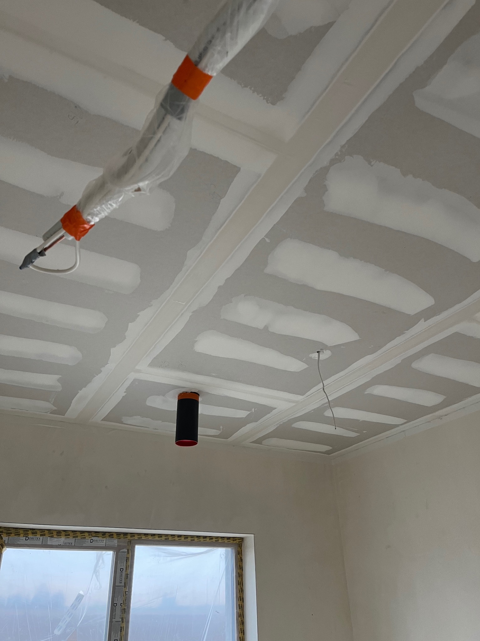 Sufit z płyt gipsowo-kartonowych w trakcie montażu, widoczne łączenia i instalacje zabezpieczone folią, okno z folią ochronną. Jasne wnętrze w trakcie prac.