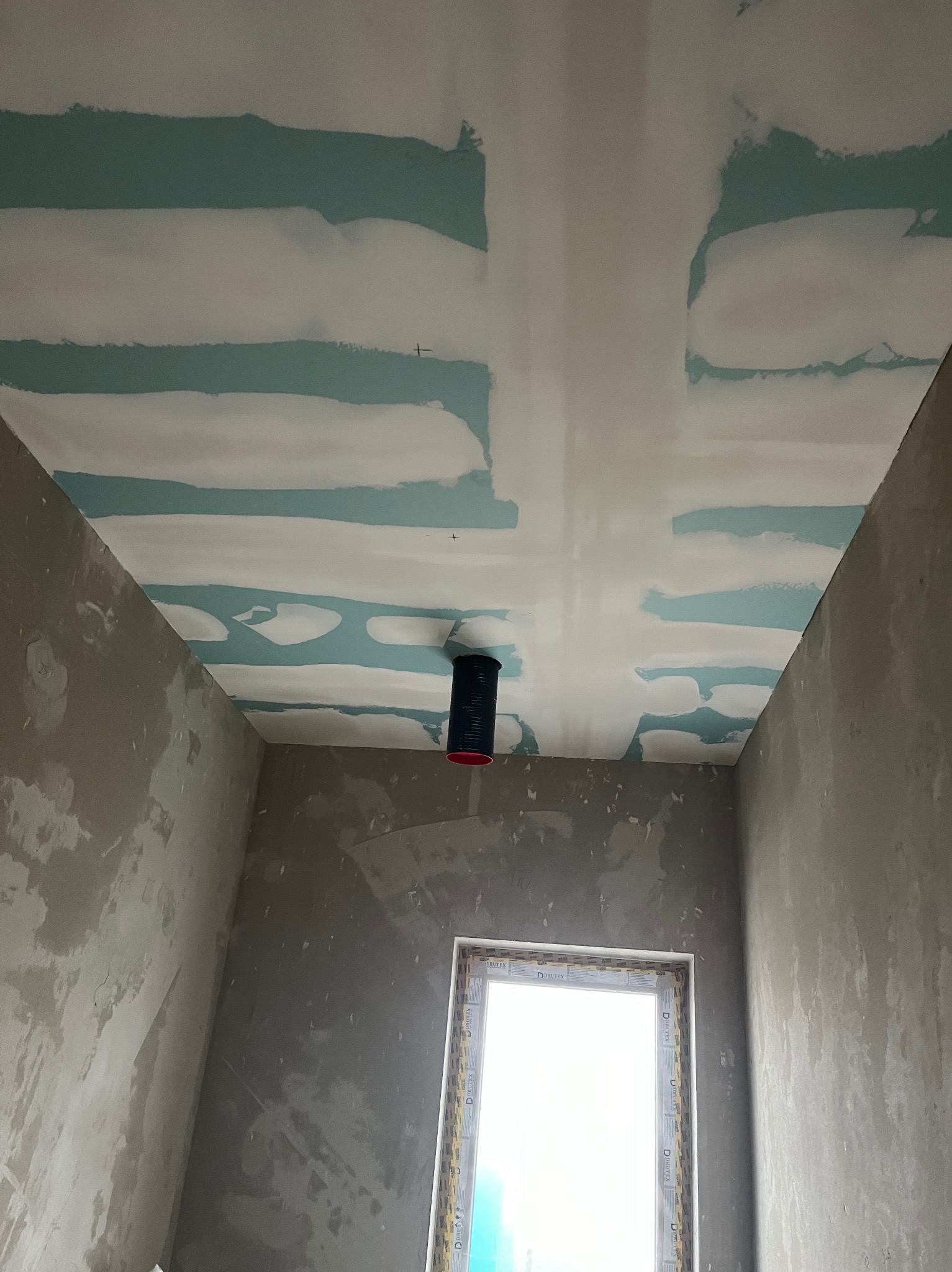 Narożne wnętrze w trakcie prac wykończeniowych: sufit z płyt gipsowo-kartonowych z widocznymi łączeniami, ściany w szarym tynku, okno z PCV.