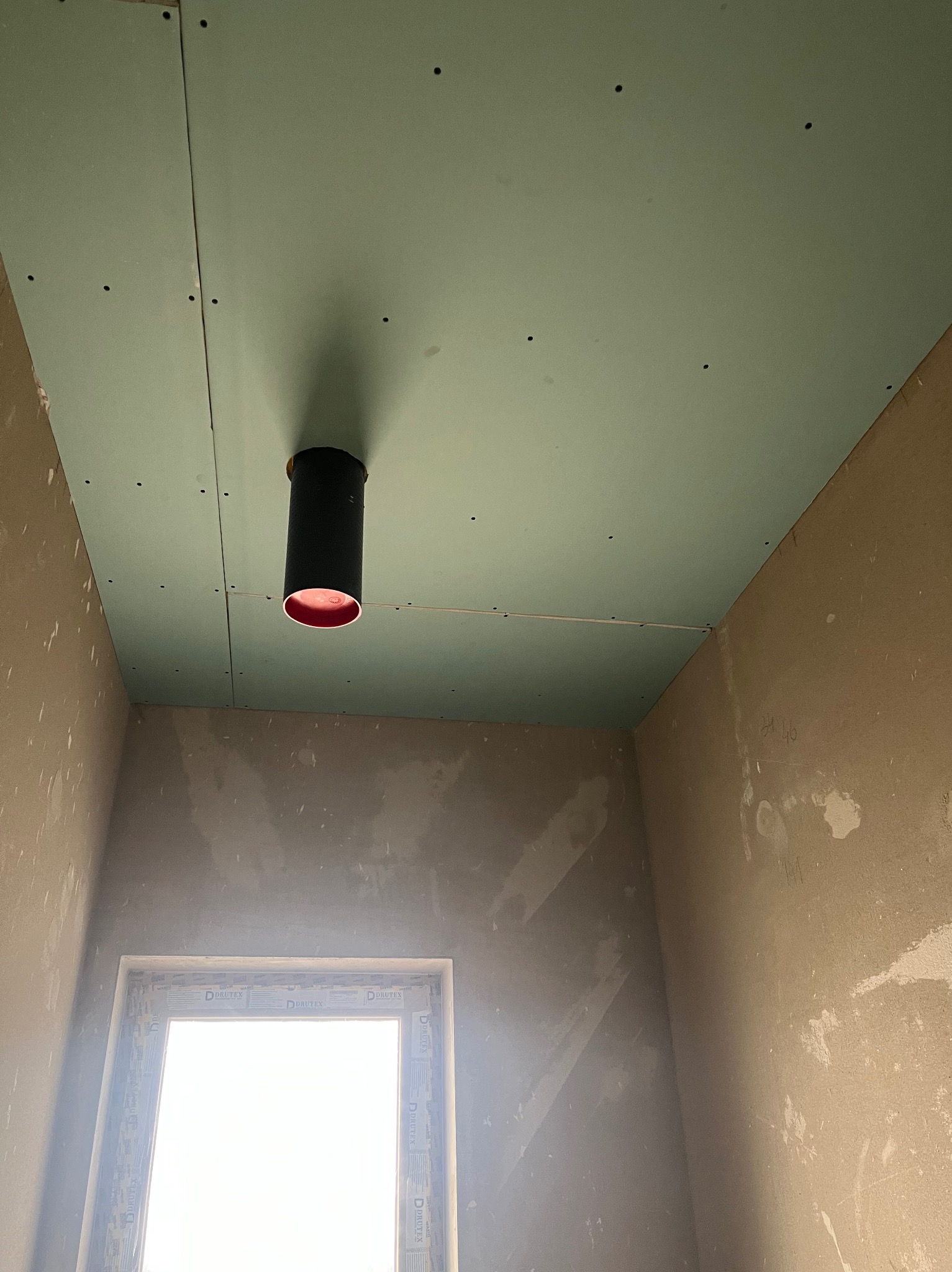 Montaż płyty G-K na suficie z widocznym otworem wentylacyjnym. Ściany w stanie surowym, okno z widocznym profilem. Ujęcie z dołu, podkreślające etap prac.