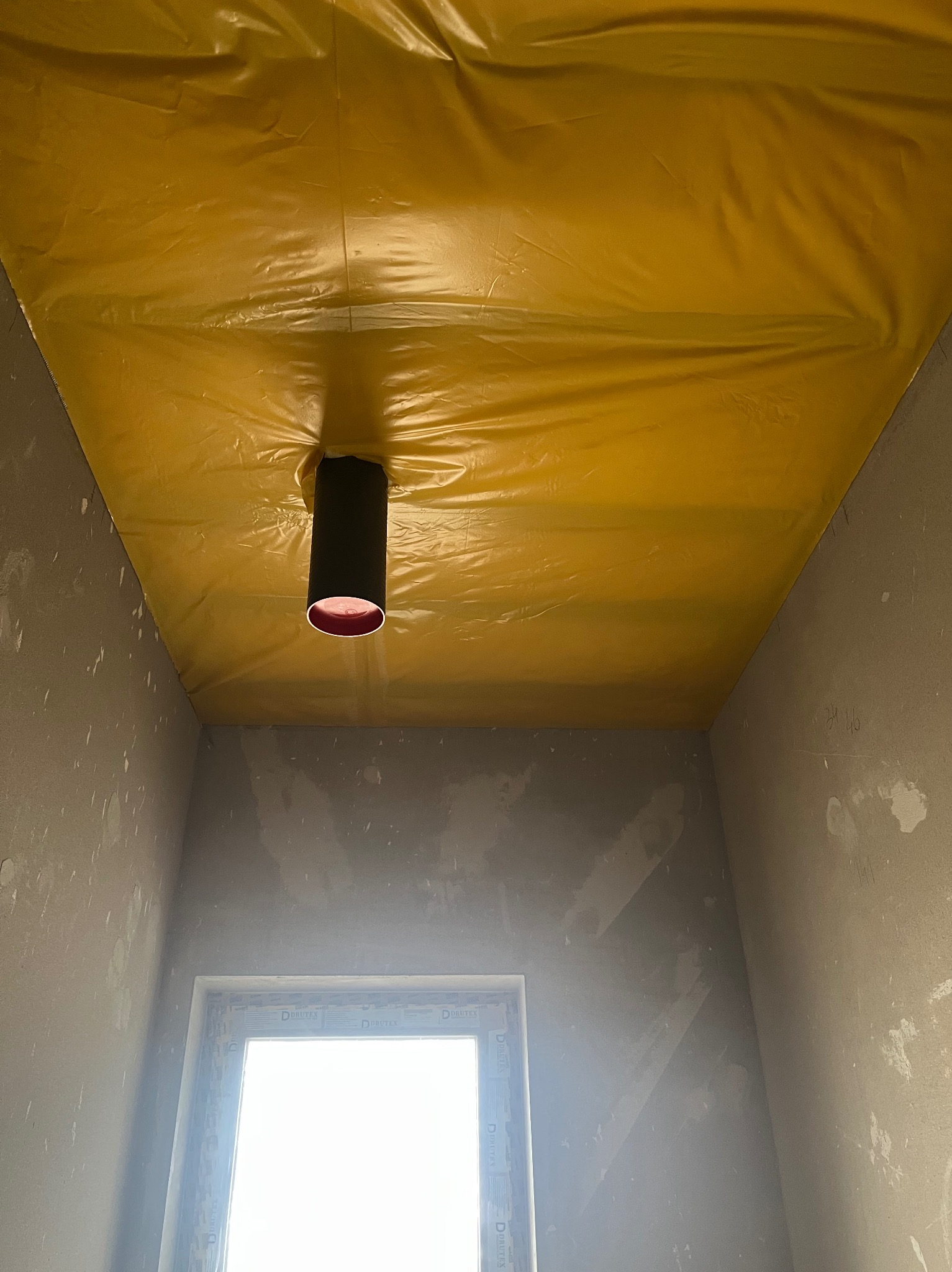 Żółta folia zabezpieczająca sufit podczas prac wykończeniowych, z widocznym otworem na lampę. Ściany w stanie surowym, okno w tle. Przygotowanie do dalszych prac.