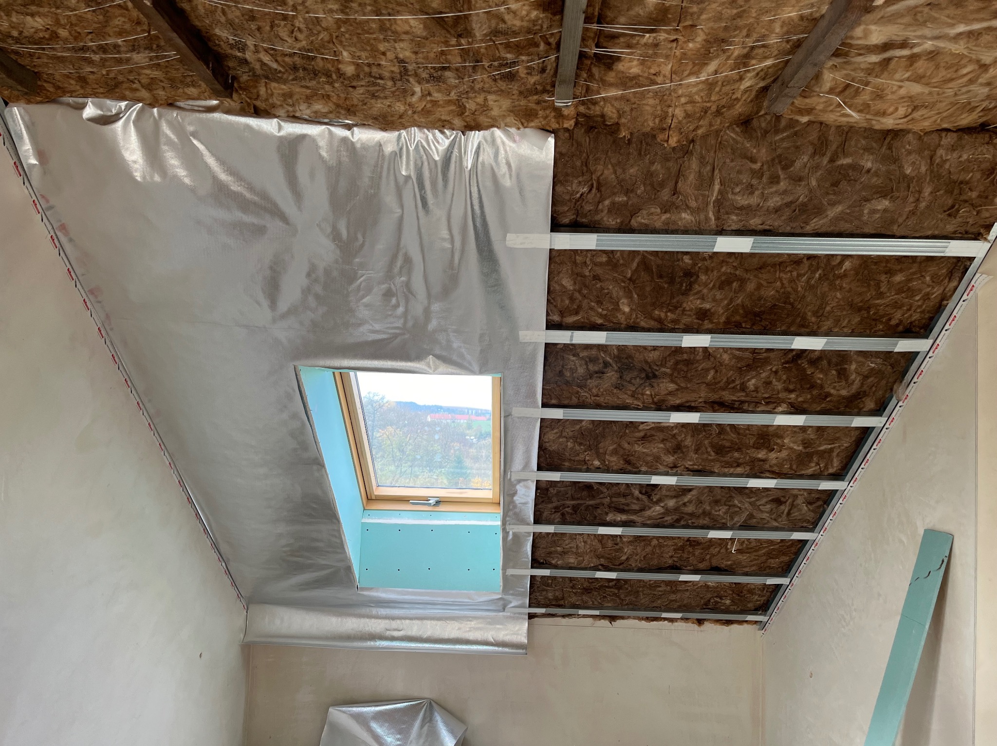 Skośny sufit poddasza w trakcie izolacji: wełna mineralna, folia aluminiowa i metalowy stelaż. Widoczne okno dachowe i fragment ściany. Prace wykończeniowe.