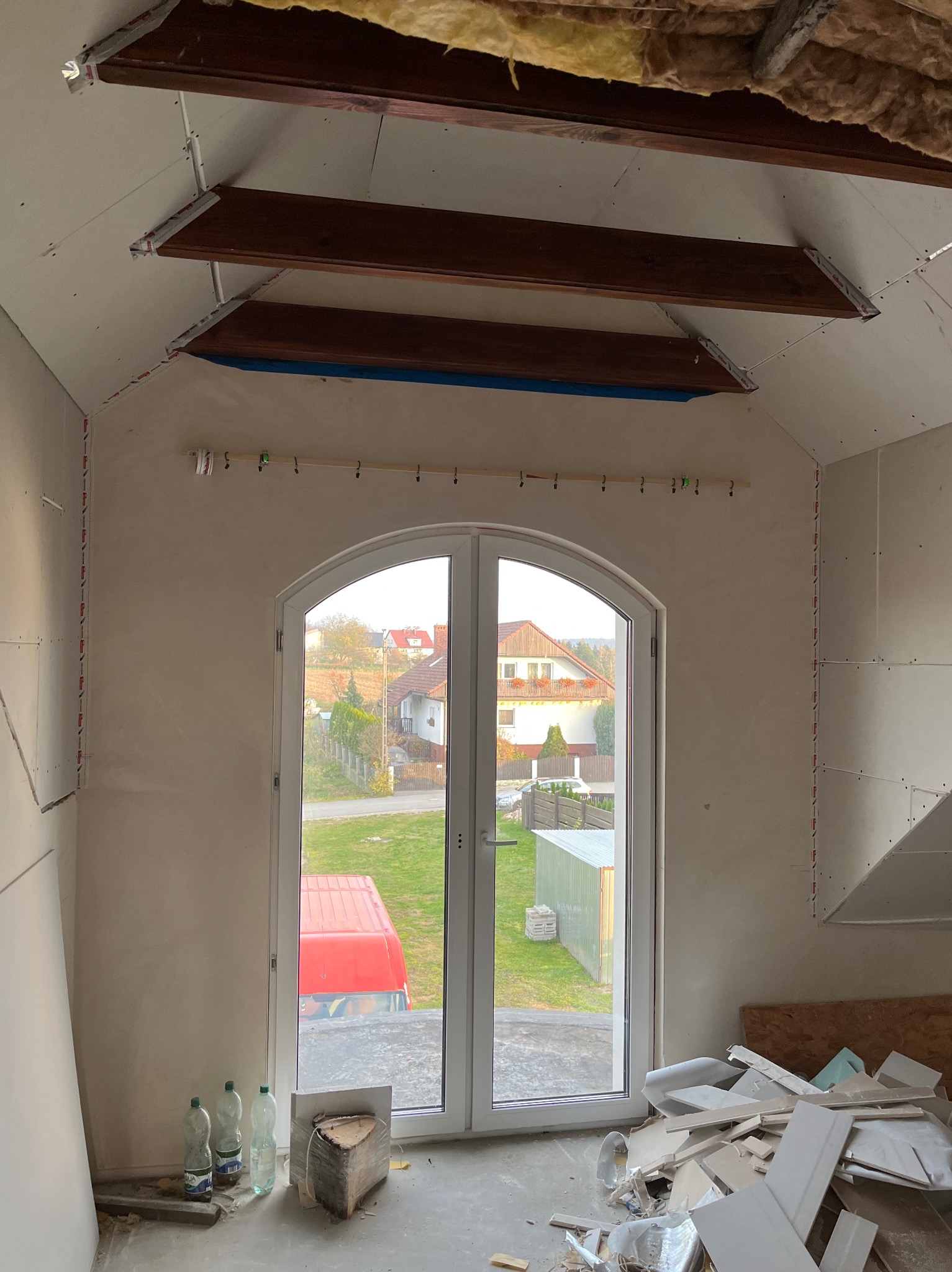 Poddasze w trakcie adaptacji: widoczne płyty gipsowo-kartonowe na ścianach i suficie, drewniane belki konstrukcyjne, okno balkonowe z widokiem na zewnątrz. Prace wykończeniowe w toku.
