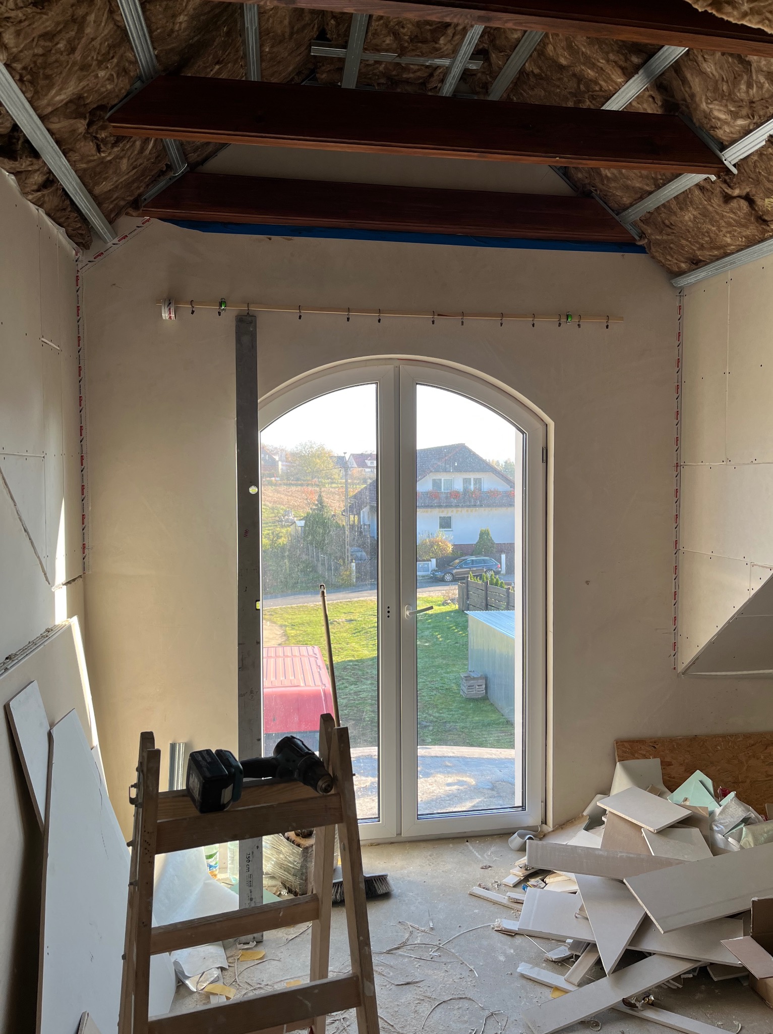 Poddasze w trakcie prac wykończeniowych: ocieplenie wełną, montaż płyt gipsowych, widoczne narzędzia i resztki materiałów. Okno z widokiem na zewnątrz.