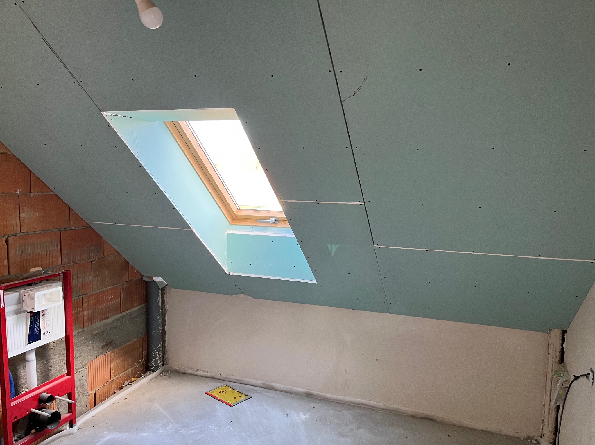 Poddasze z oknem dachowym w trakcie montażu płyt gipsowo-kartonowych. Widoczna instalacja sanitarna przy ceglanej ścianie i surowy beton na podłodze.