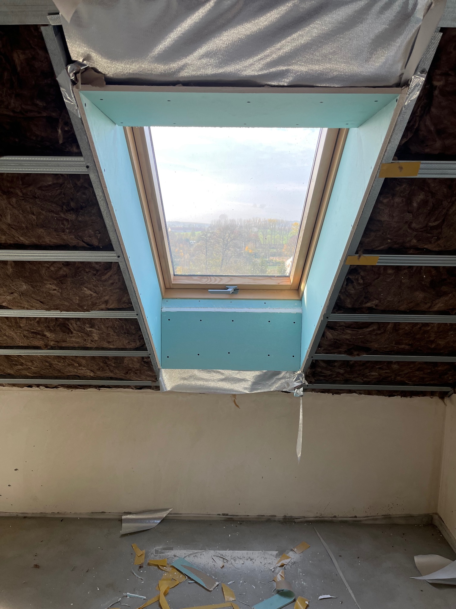 Montaż okna dachowego z płytą GK na poddaszu. Widoczne ocieplenie wełną mineralną i metalowy stelaż. Fragment ściany z widocznymi śladami prac wykończeniowych.