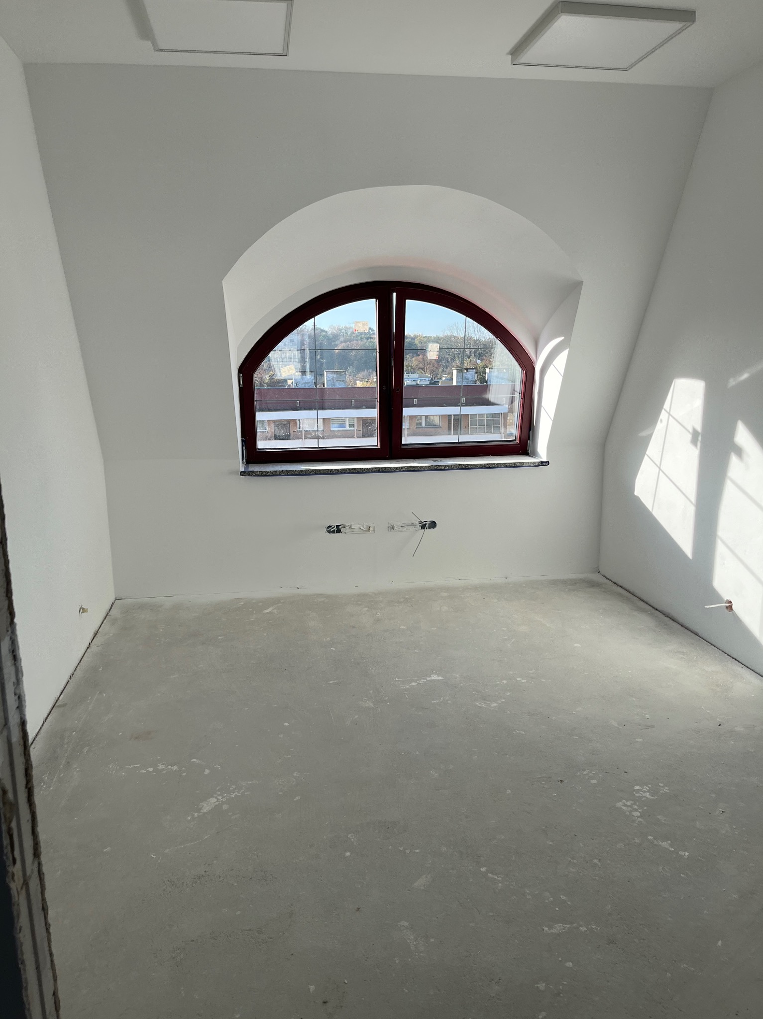 Poddasze po remoncie: białe ściany, szara podłoga, okno łukowe z ciemną ramą. Widoczne wystające kable elektryczne. Dwa kwadratowe panele oświetleniowe na suficie.