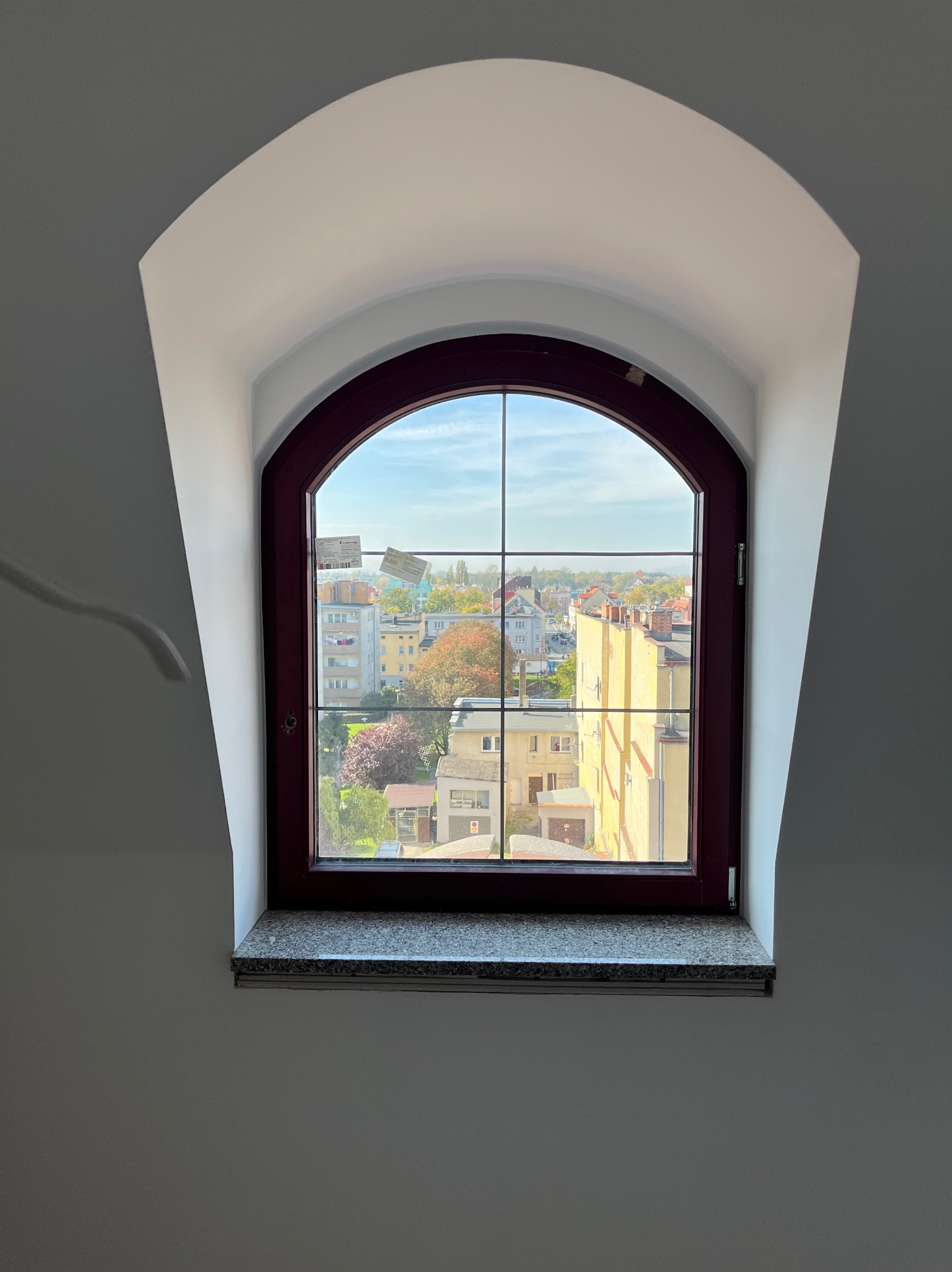 Arched window with dark wood frame, divided panes, and a view of a cityscape with autumn foliage. White walls and a granite windowsill complete the interior.