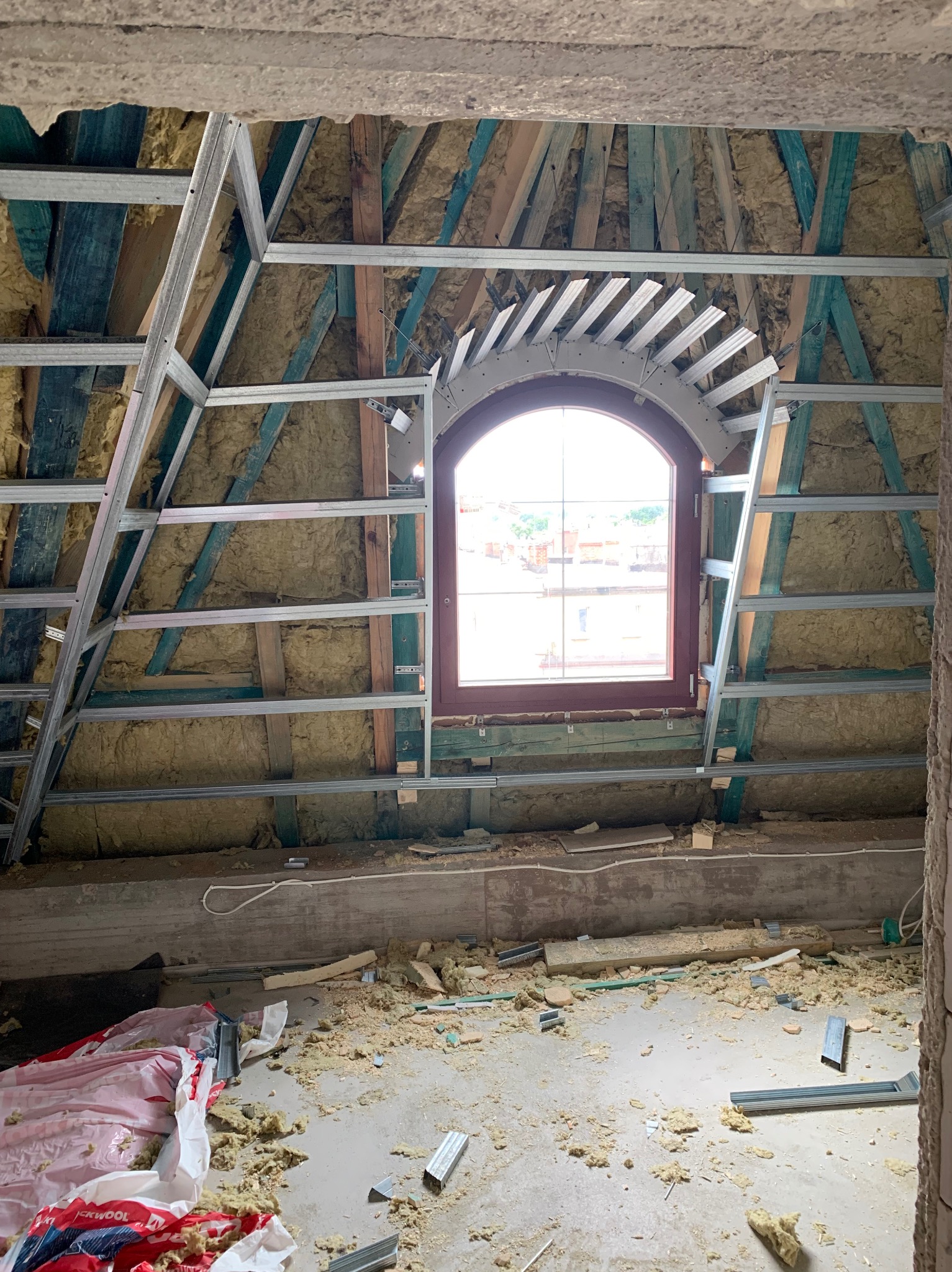 Poddasze w trakcie izolacji: wełna mineralna między krokwiami, metalowy stelaż pod płytę G-K, okno łukowe. Widoczne resztki materiałów na podłodze.