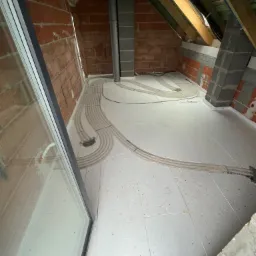 Podłogowe ogrzewanie rekuperacyjne w trakcie instalacji na poddaszu, widoczne rury rozprowadzone na izolacji podłogowej, ceglana ściana w tle.