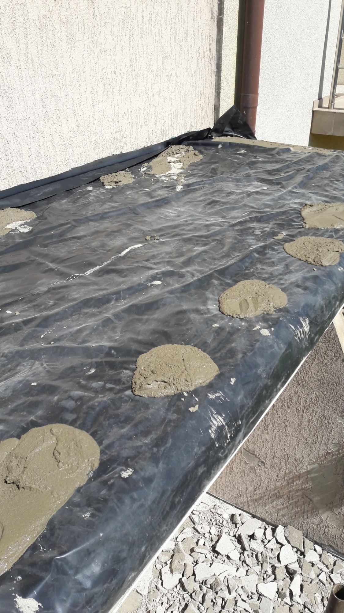 Przygotowanie do prac elewacyjnych: czarna folia zabezpieczająca i porcje zaprawy murarskiej na podłożu, widoczne fragmenty ściany z fakturą.