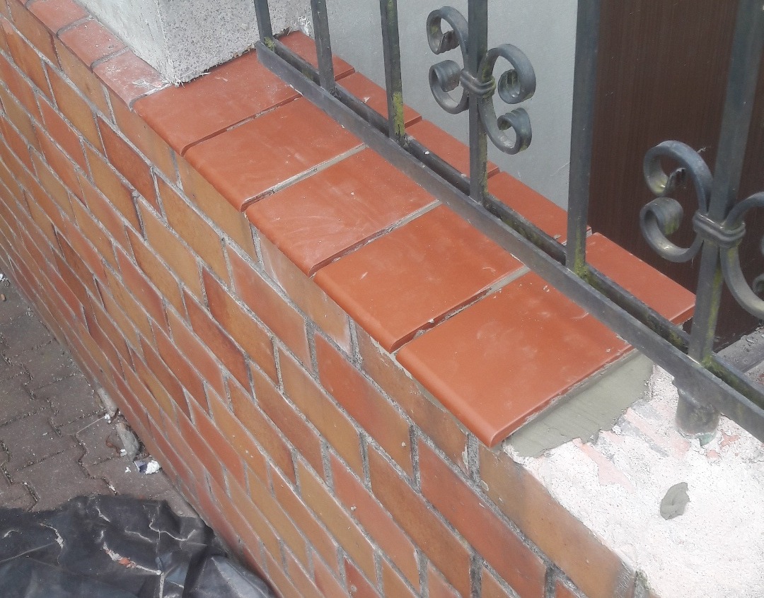 Murowana ścianka z cegły klinkierowej, wykończona ceramicznymi płytkami na górze, z metalowym ozdobnym ogrodzeniem. Widoczne ślady zaprawy i pozostałości po pracach budowlanych.