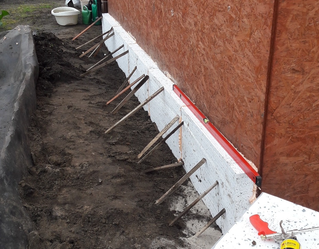 Ocieplanie fundamentów styropianem z użyciem rozpórek i poziomicy, widoczna folia hydroizolacyjna i pistolet do pianki montażowej.