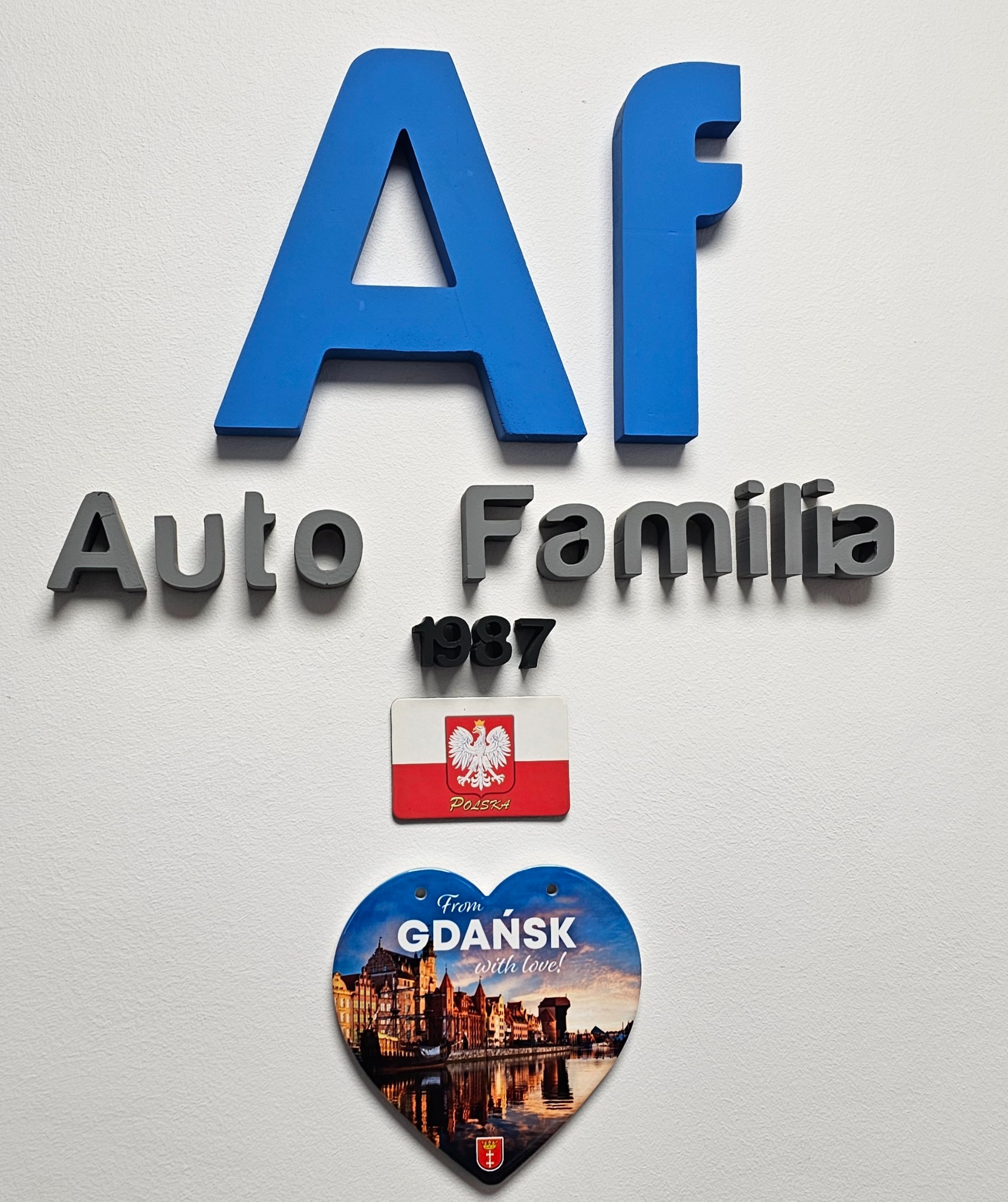 Ściana z logo firmy Auto Familia z Gdańska, rok założenia 1987, ozdobiona magnesem z flagą Polski i magnesem w kształcie serca z panoramą Gdańska.