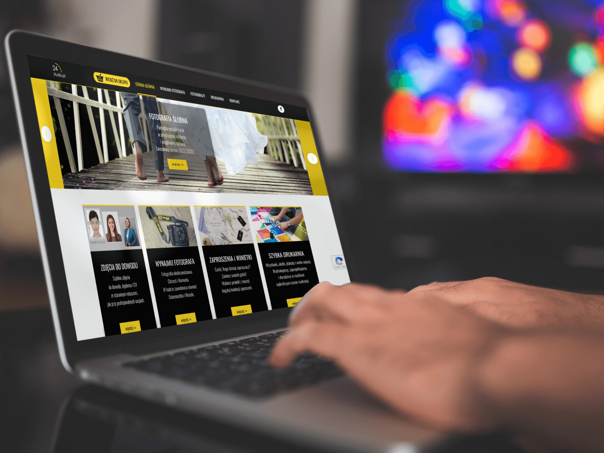 Osoba pisze na laptopie, na ekranie strona internetowa fotografa ślubnego z ofertą zdjęć do dowodu, wynajmu fotografa, zaproszeń i wizytówek oraz szybkiej drukarni, w tle rozmyte kolorowe światła.