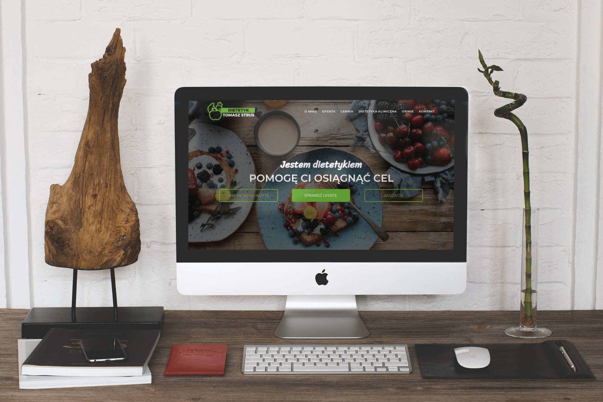Ekran komputera iMac wyświetlający stronę internetową dietetyka Tomasza Strusa, z drewnianą rzeźbą, bambusem w wazonie, notatnikami i akcesoriami na drewnianym biurku.