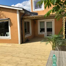 Jasna drewniana deska tarasowa przed pomarańczowym domem z oknami i zadaszeniem, widoczne meble ogrodowe i roślina doniczkowa.