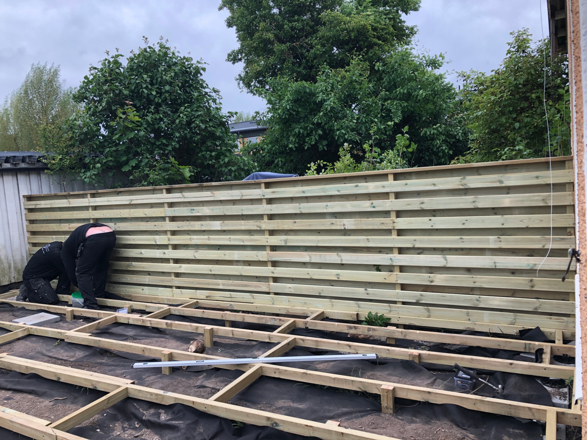 Budowa drewnianego tarasu: pracownicy montują legary na czarnej geowłókninie, w tle świeżo postawione ogrodzenie z poziomych desek.