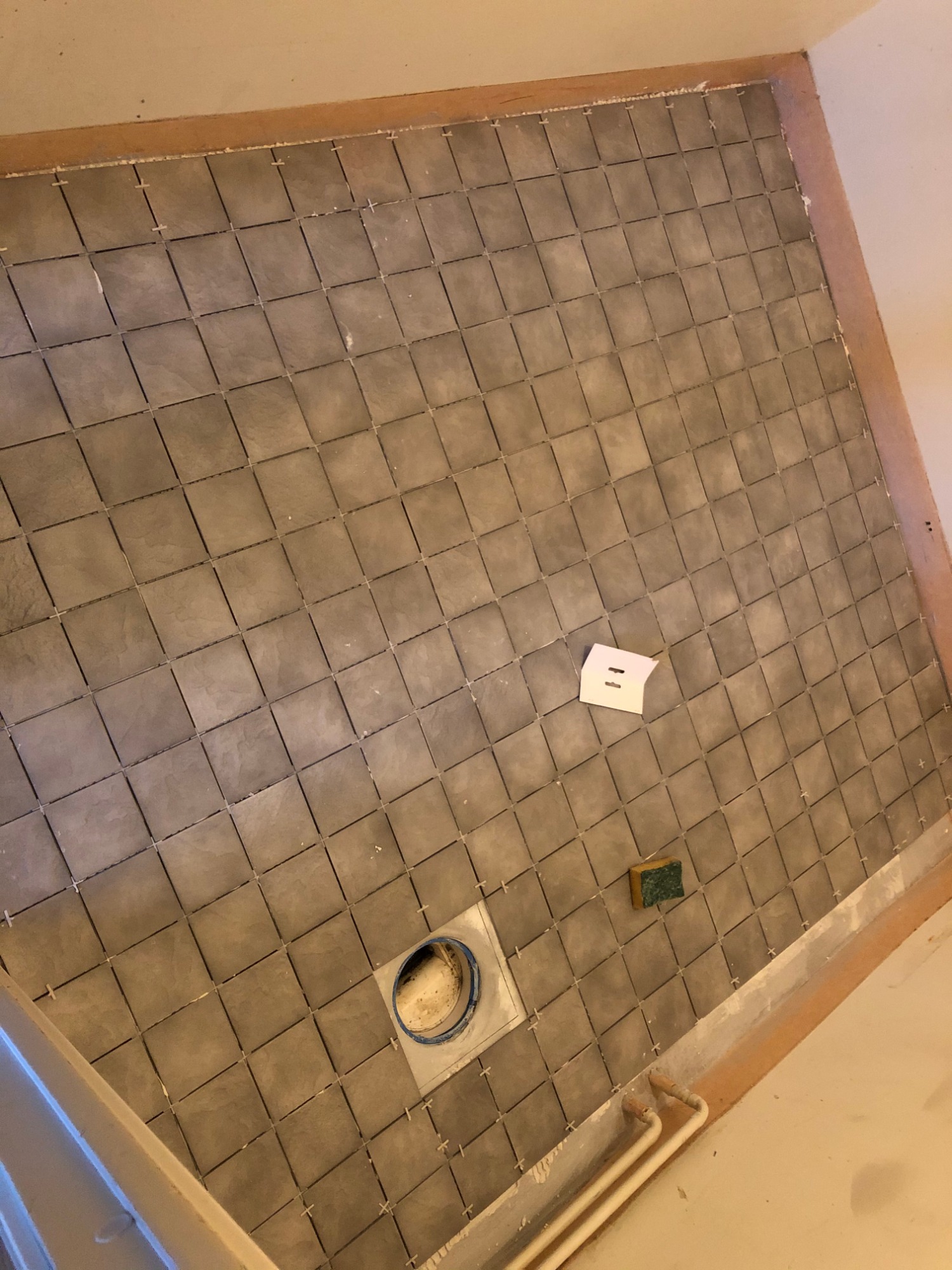 Układanie szarych, kwadratowych płytek w łazience, widoczny odpływ liniowy, krzyżyki dystansowe, gąbka i szablon.