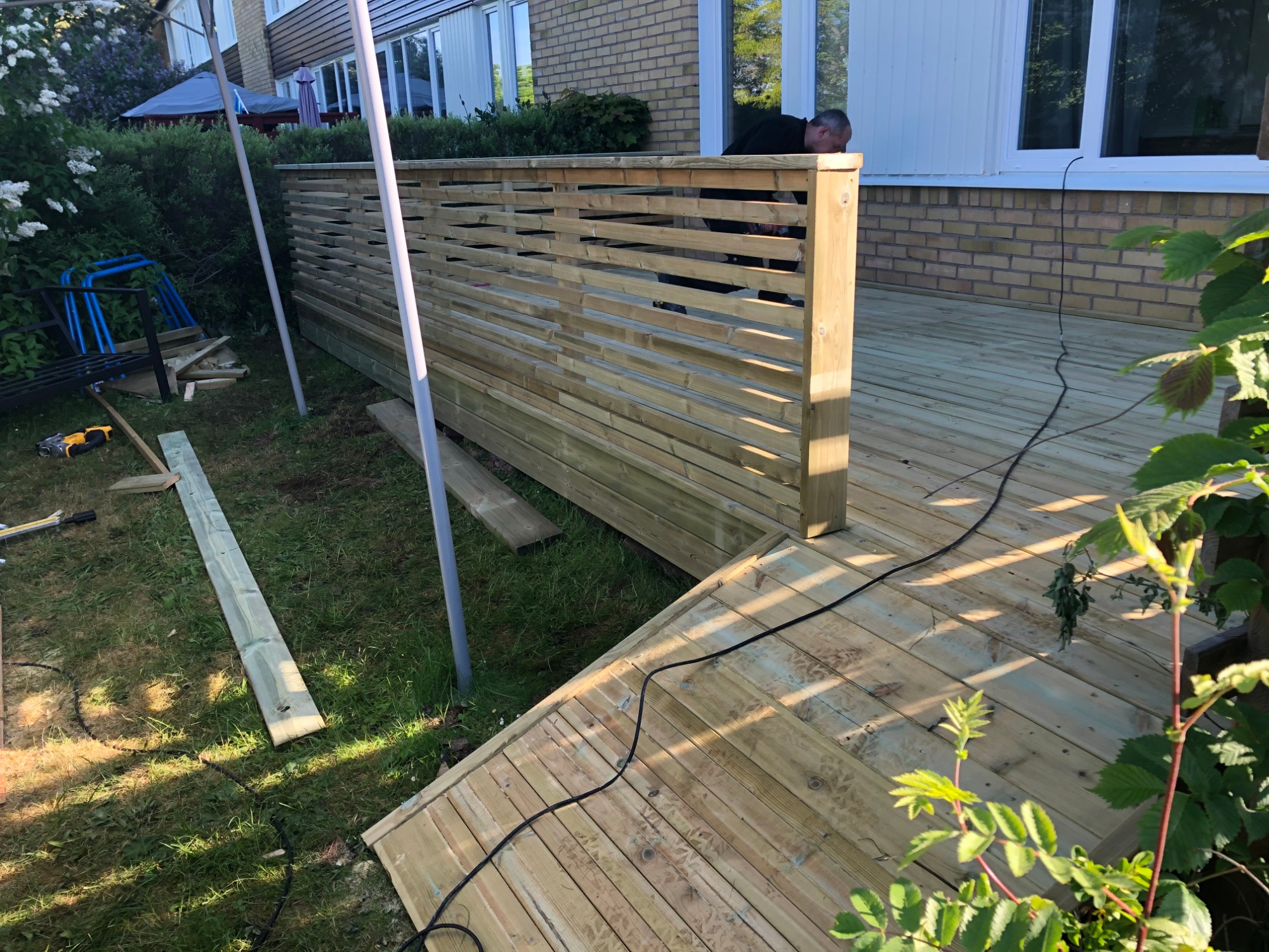 Montaż drewnianego tarasu z ażurową balustradą przy domu z cegły, widoczne narzędzia i elementy konstrukcyjne na trawie, pracownik w tle.